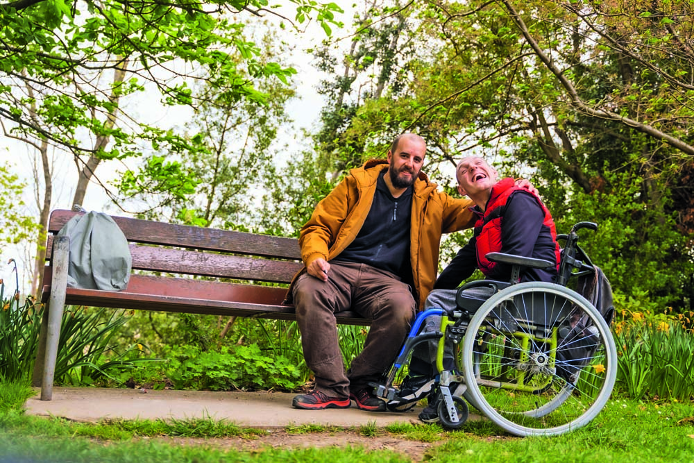 Portrait of a paralyzed young man in a wheelchair with a friend on a bench in a public park in the city